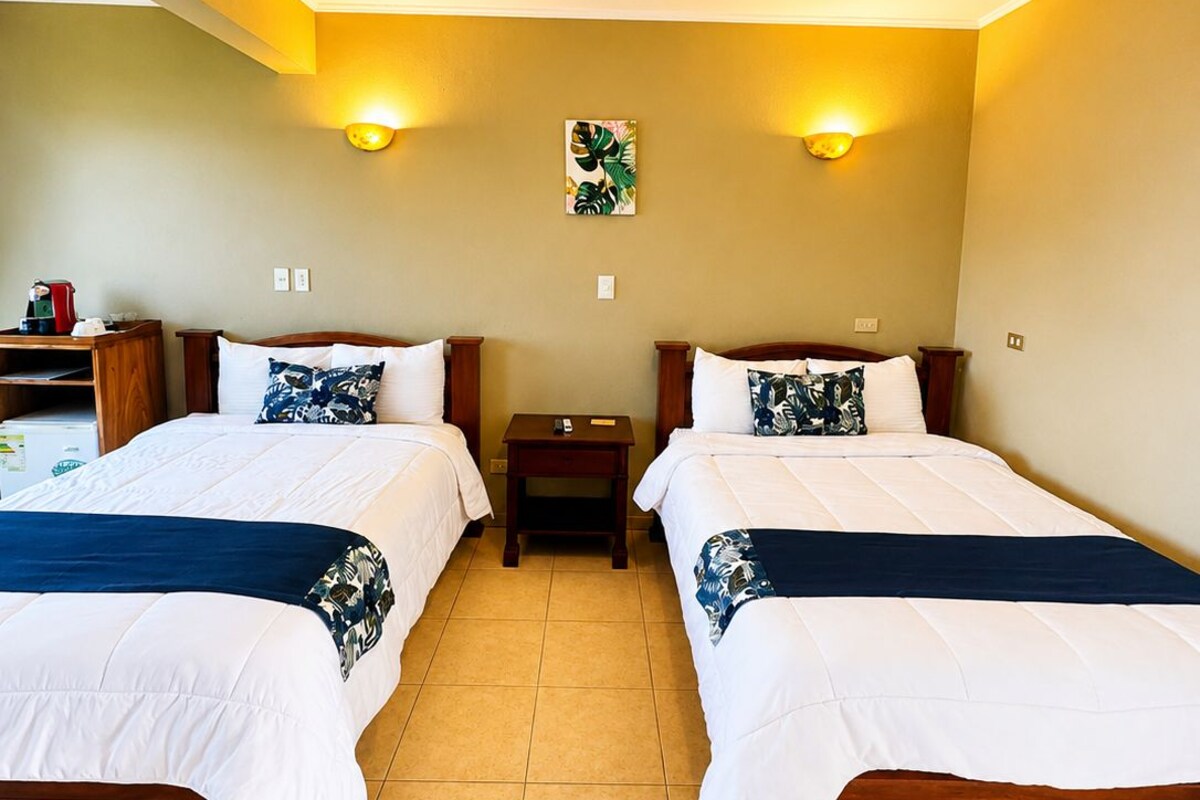 A hotel room with two beds, a nightstand, a wall-mounted light, and a painting.