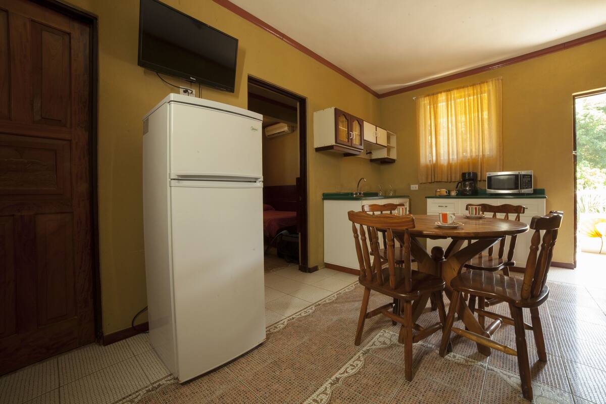 A compact kitchen with a refrigerator, wooden chairs, a dining table, a microwave, and a window with curtains.