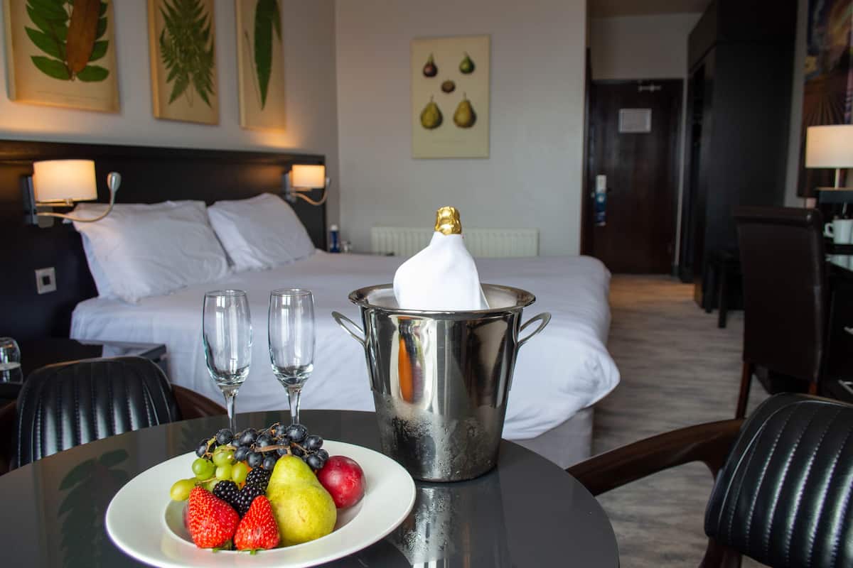 A hotel room with a bed, a dining table with fruit and champagne, and a view of the room.