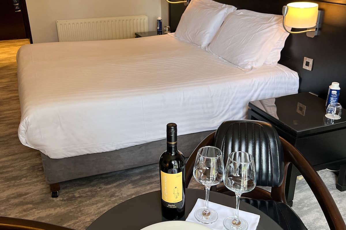 A hotel room with a large bed, a bedside table with a bottle of wine and two glasses, a plate with macarons and a chocolate bar, and wall art depicting pears and leaves.