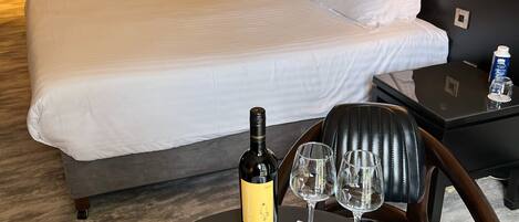 Una habitación de hotel con una cama grande, una mesita de noche con una botella de vino y dos copas, un plato con macarons y una barra de chocolate, y una obra de arte en la pared que muestra peras y hojas.