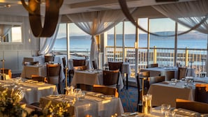 Un restaurante con mesas dispuestas para comer, amplias ventanas con vista al mar y cortinas elegantes.
