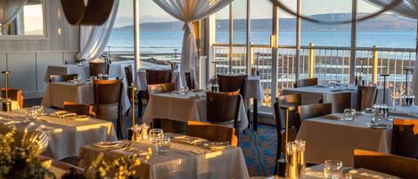 Un restaurante con mesas dispuestas para comer, amplias ventanas con vista al mar y cortinas elegantes.