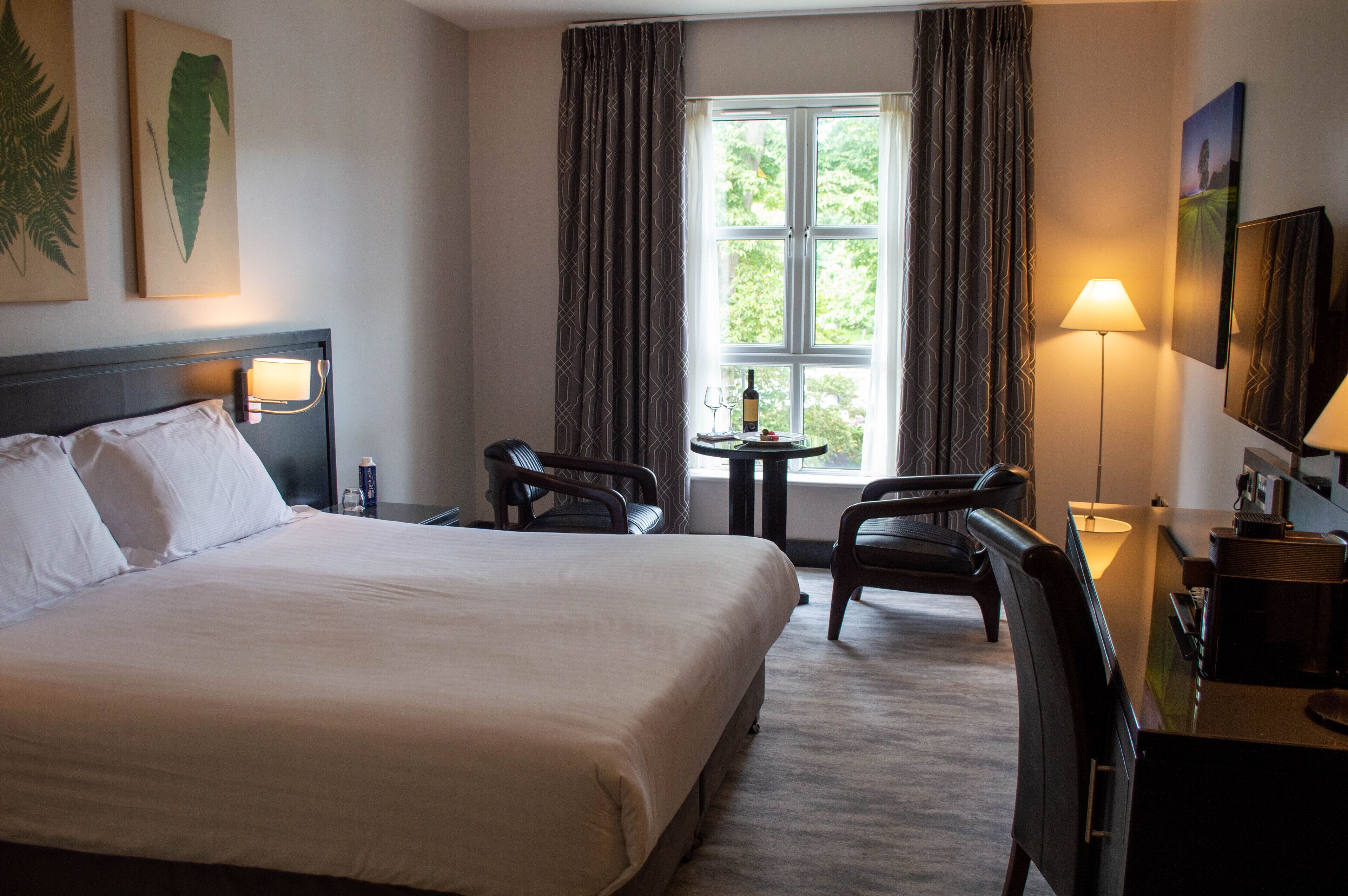 A hotel room with a large bed, two chairs, a small table with a bottle and glass, and a window with curtains.