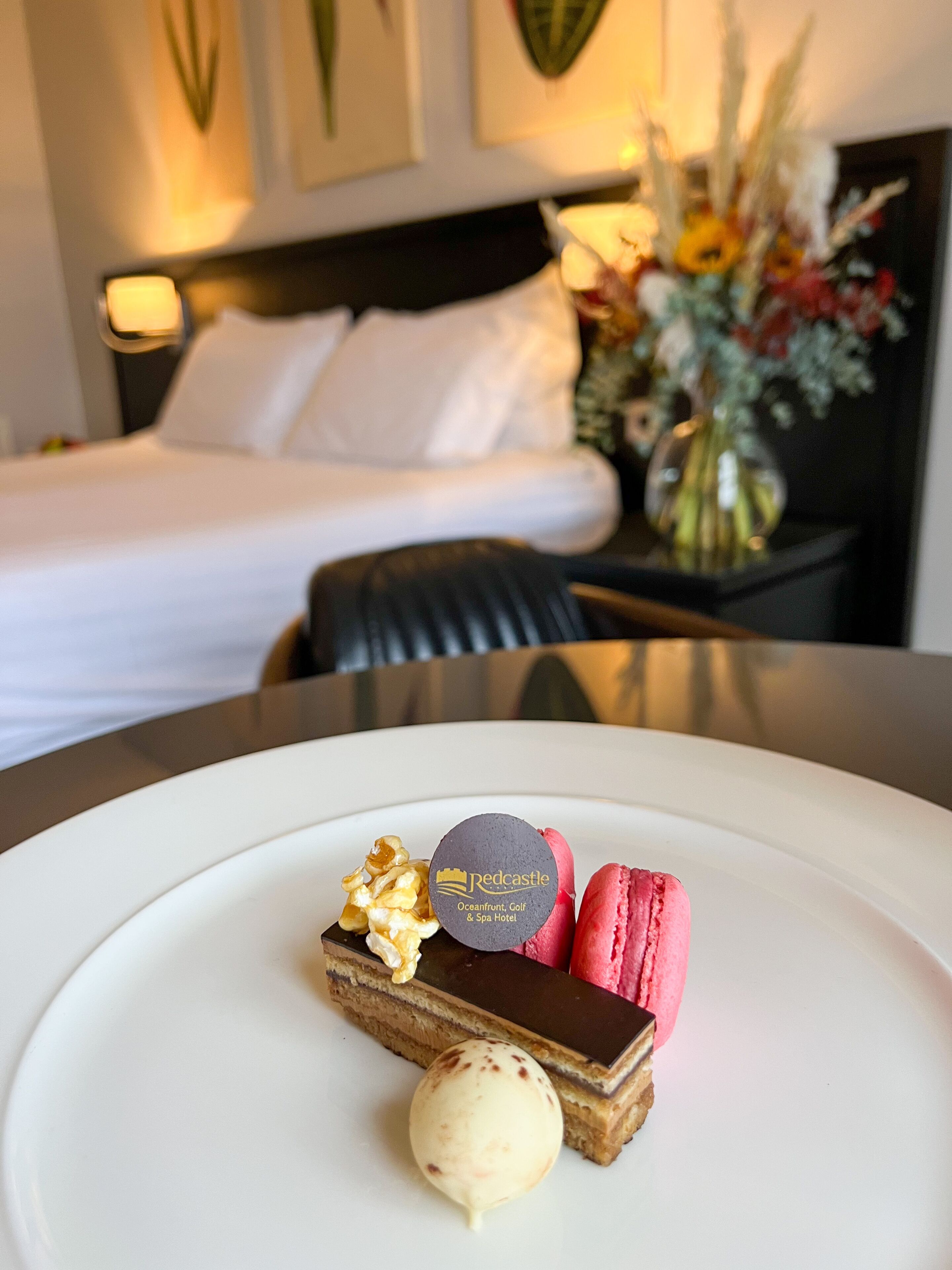 Ein Dessertteller mit Schokoladentarte, Macarons und einer Kugel Eis auf einem weißen Teller, im Hintergrund ein Bett und ein Nachttisch mit Blumen.