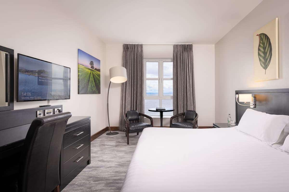 A hotel room with a bed, a desk with a television, two chairs, a table, and a window with curtains.