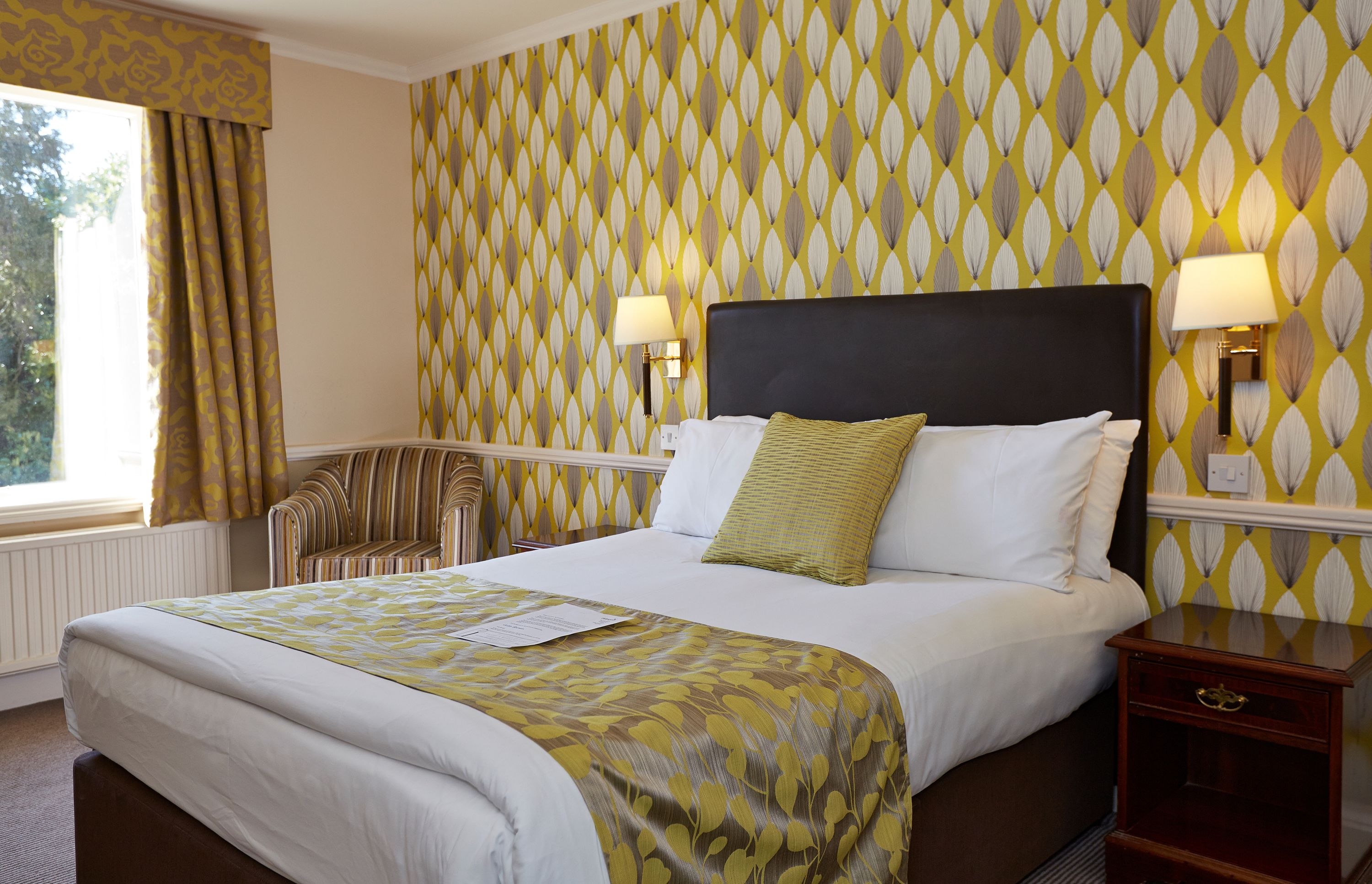 A hotel room with a bed, two bedside lamps, a nightstand, a chair, and a window with curtains.