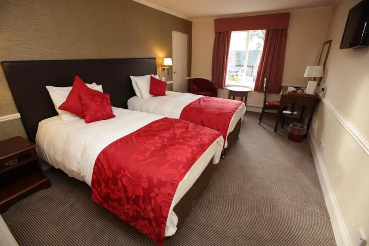 A hotel room with two beds, each with red bedding and pillows, a small desk with a chair, a television on the wall, and a window with curtains.
