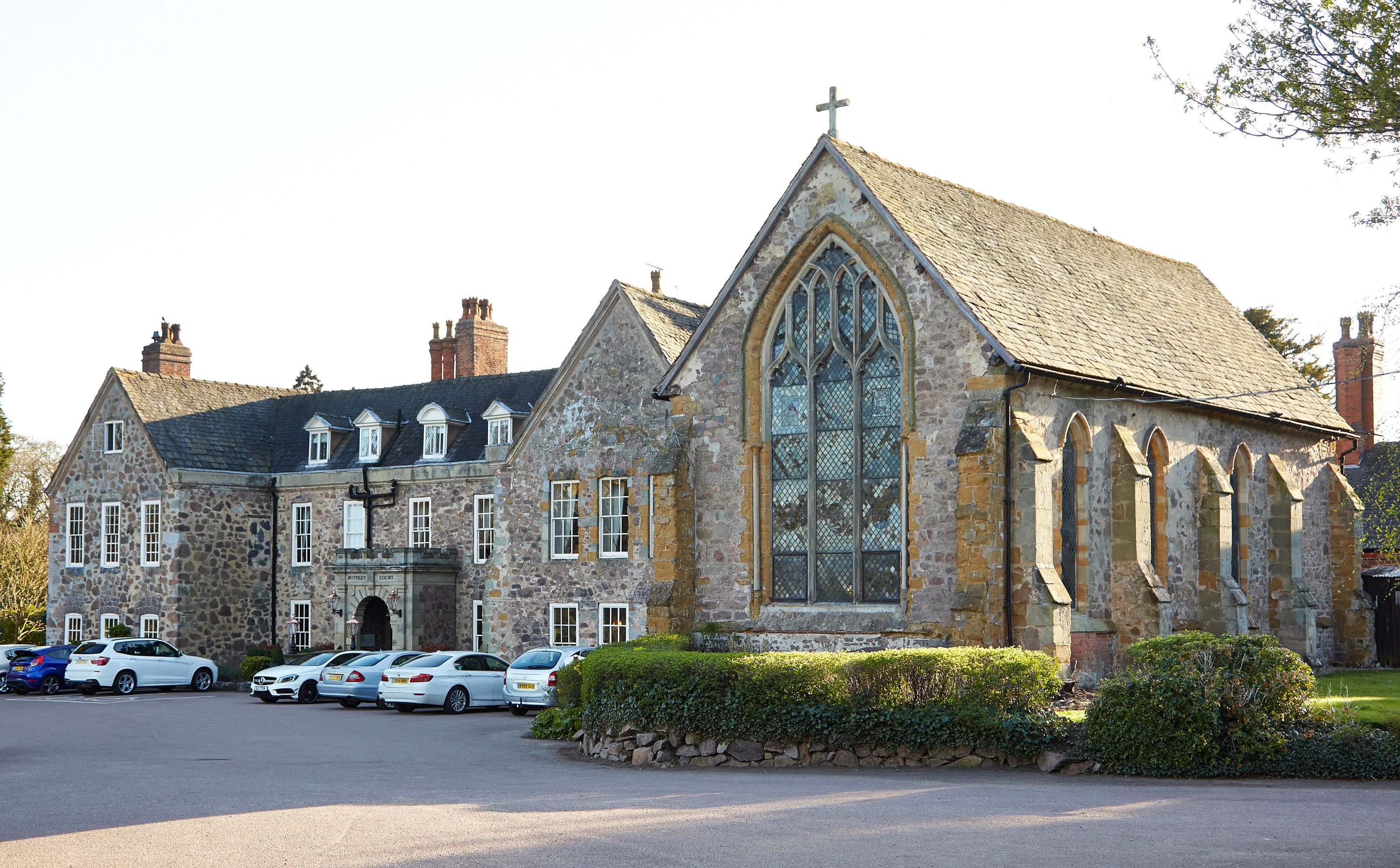A historic stone building with a large arched window and a cross on top, likely a church or chapel, with a parking lot in front.