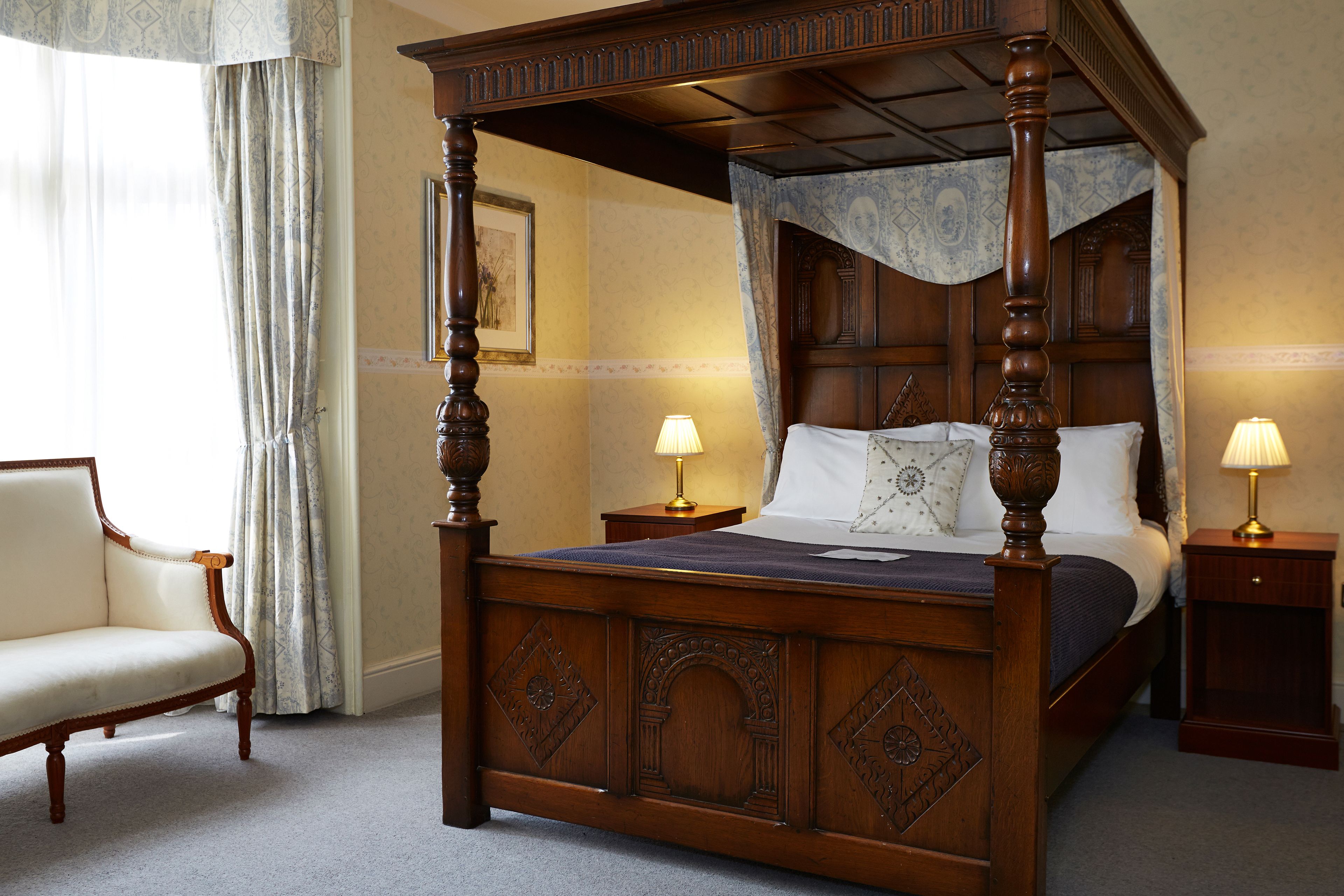 A four-poster bed with a wooden frame and a canopy, two bedside tables with lamps, a chair, and a framed picture on the wall.