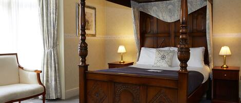 A four-poster bed with a wooden frame and a canopy, two bedside tables with lamps, a chair, and a framed picture on the wall.