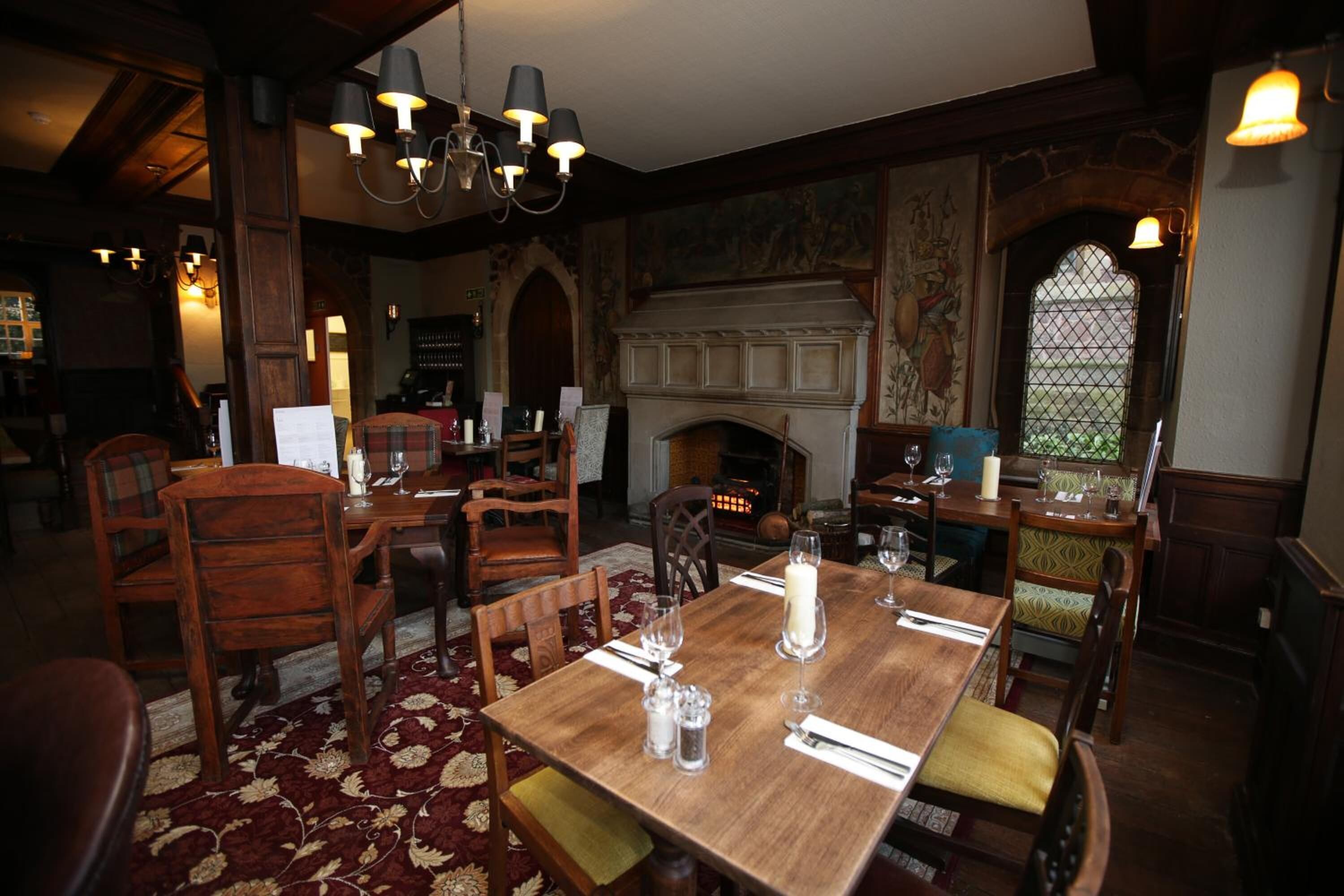 A dining area with a wooden table set for four, surrounded by wooden chairs. There is a fireplace with a stone mantle, and a large window with a decorative frame.