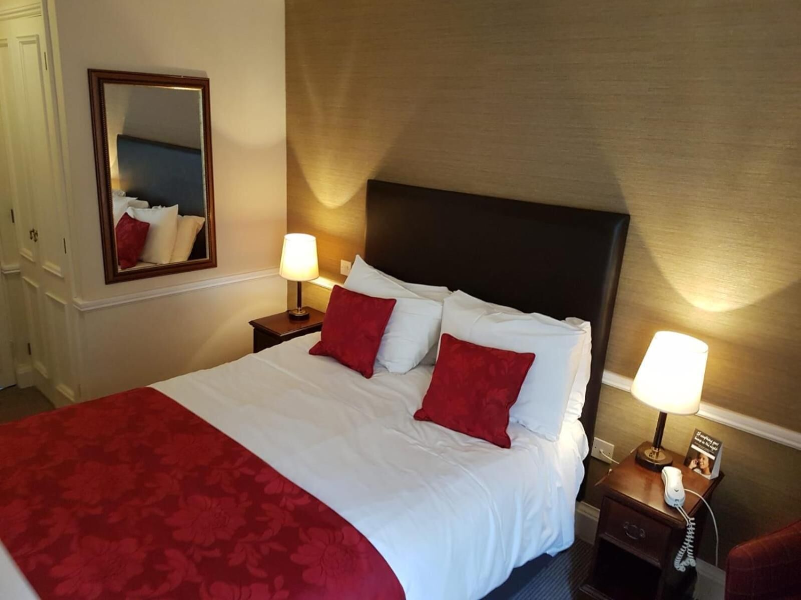 A hotel room with a large bed, two bedside lamps, a nightstand with a telephone, and a mirror on the wall.