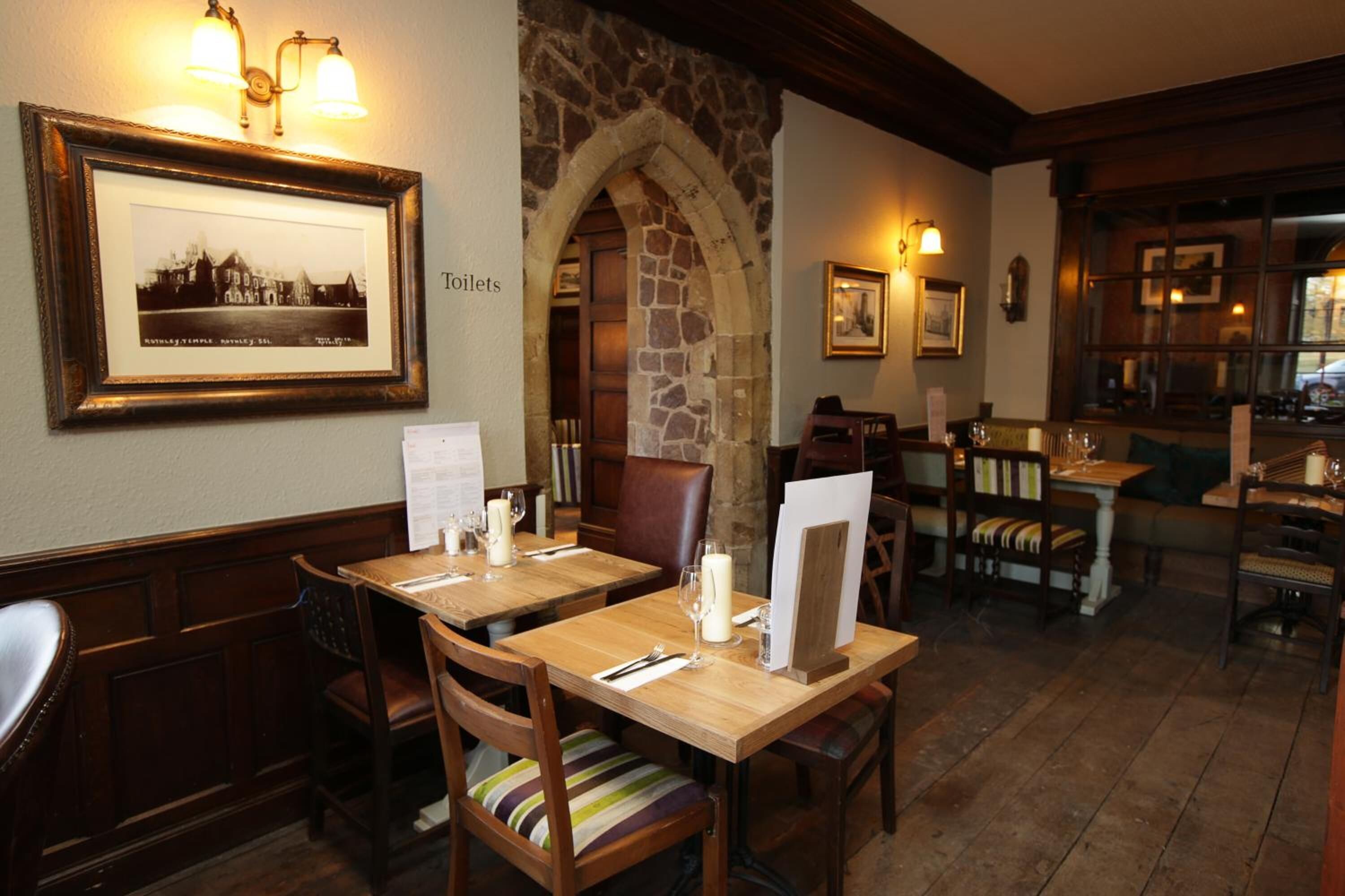 A restaurant with wooden tables and chairs, stone archway, framed picture, and a sign indicating the restroom.