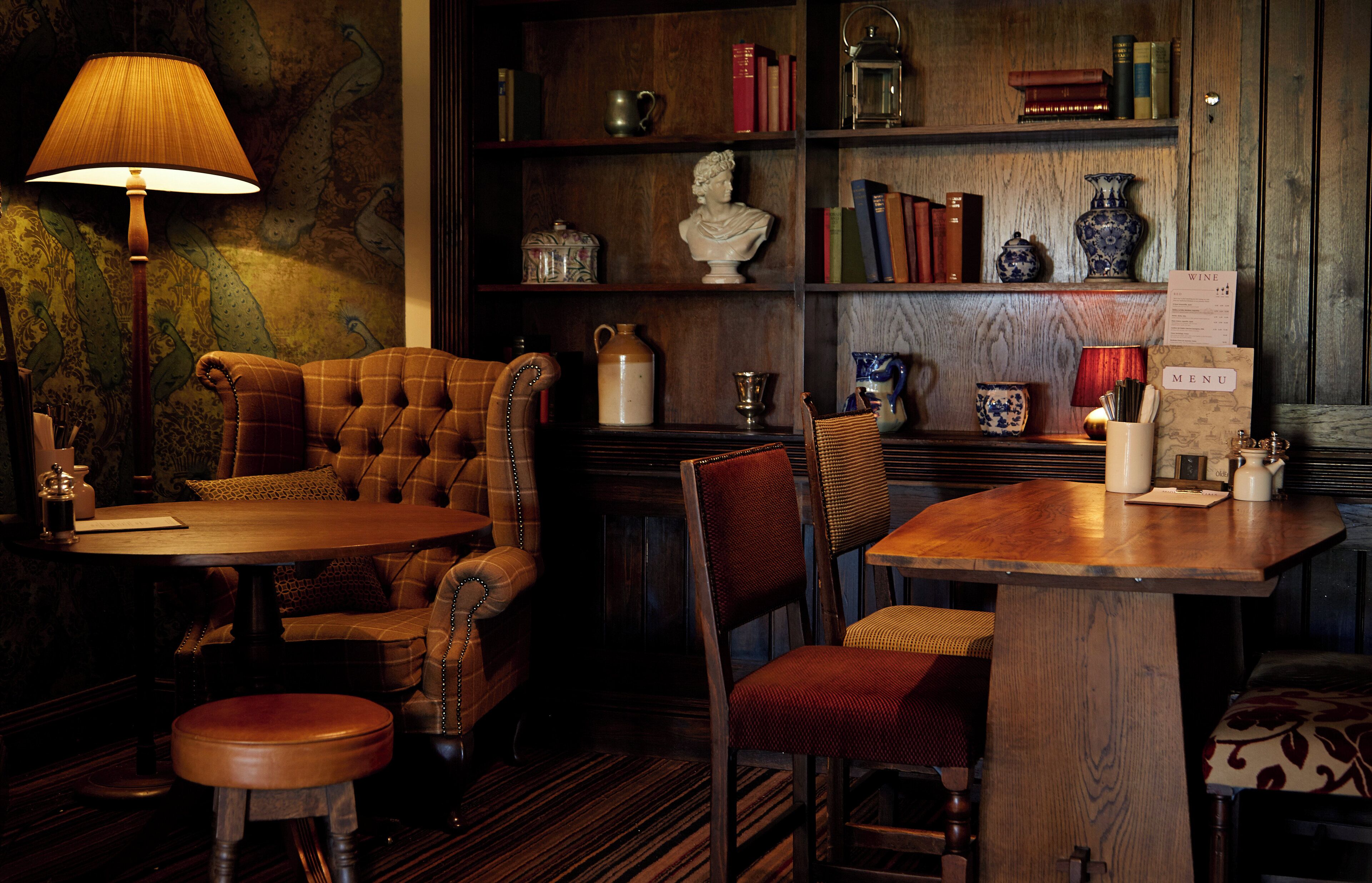 A cozy room with a wooden table, a sofa, and a chair. There is a lamp, a bookshelf with books and decorative items, and a menu sign.
