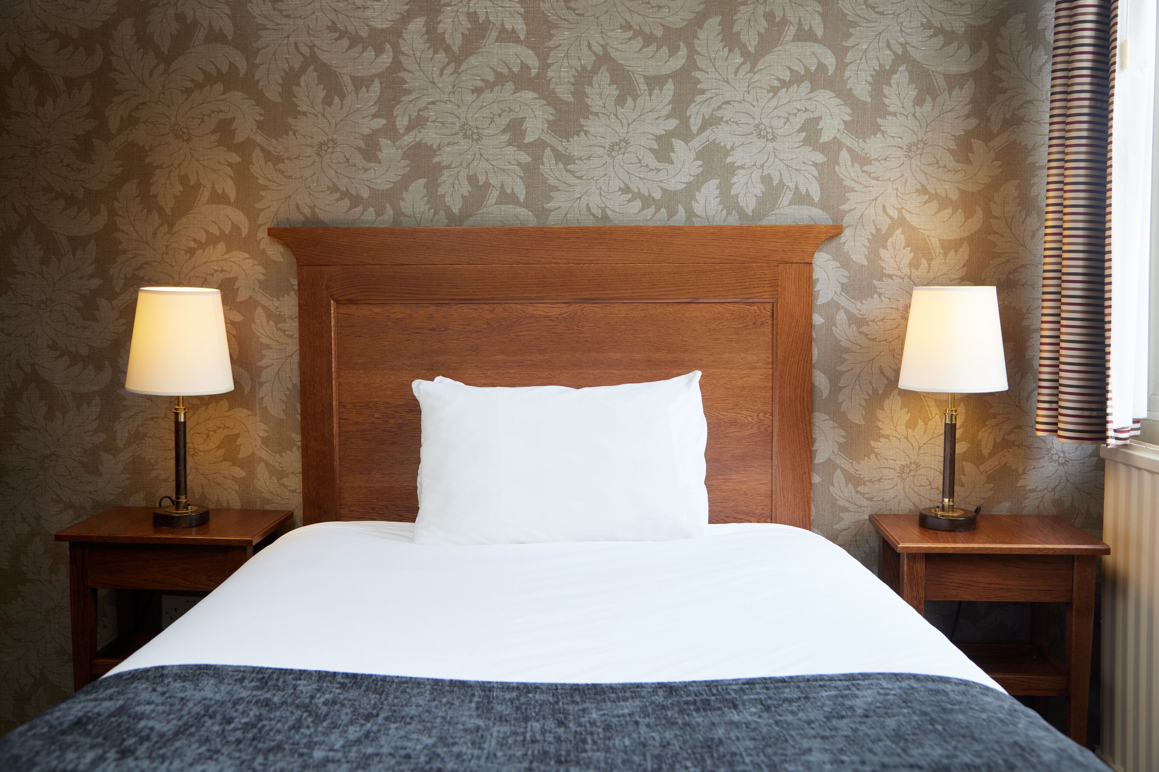 A neatly made bed with a wooden headboard, two bedside lamps, and a floral-patterned wallpaper.