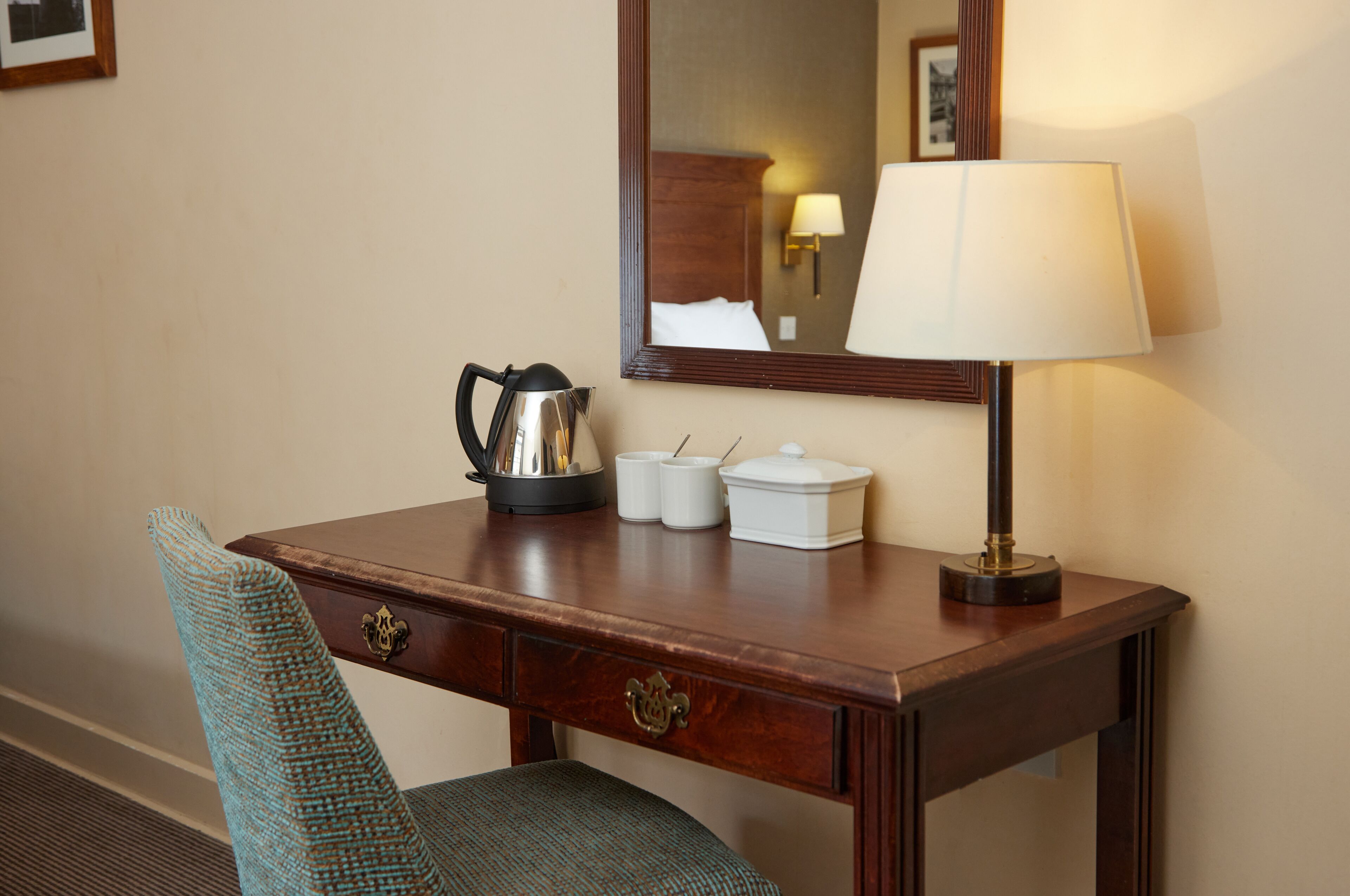 A wooden desk with a lamp, a kettle, and a chair in a hotel room.