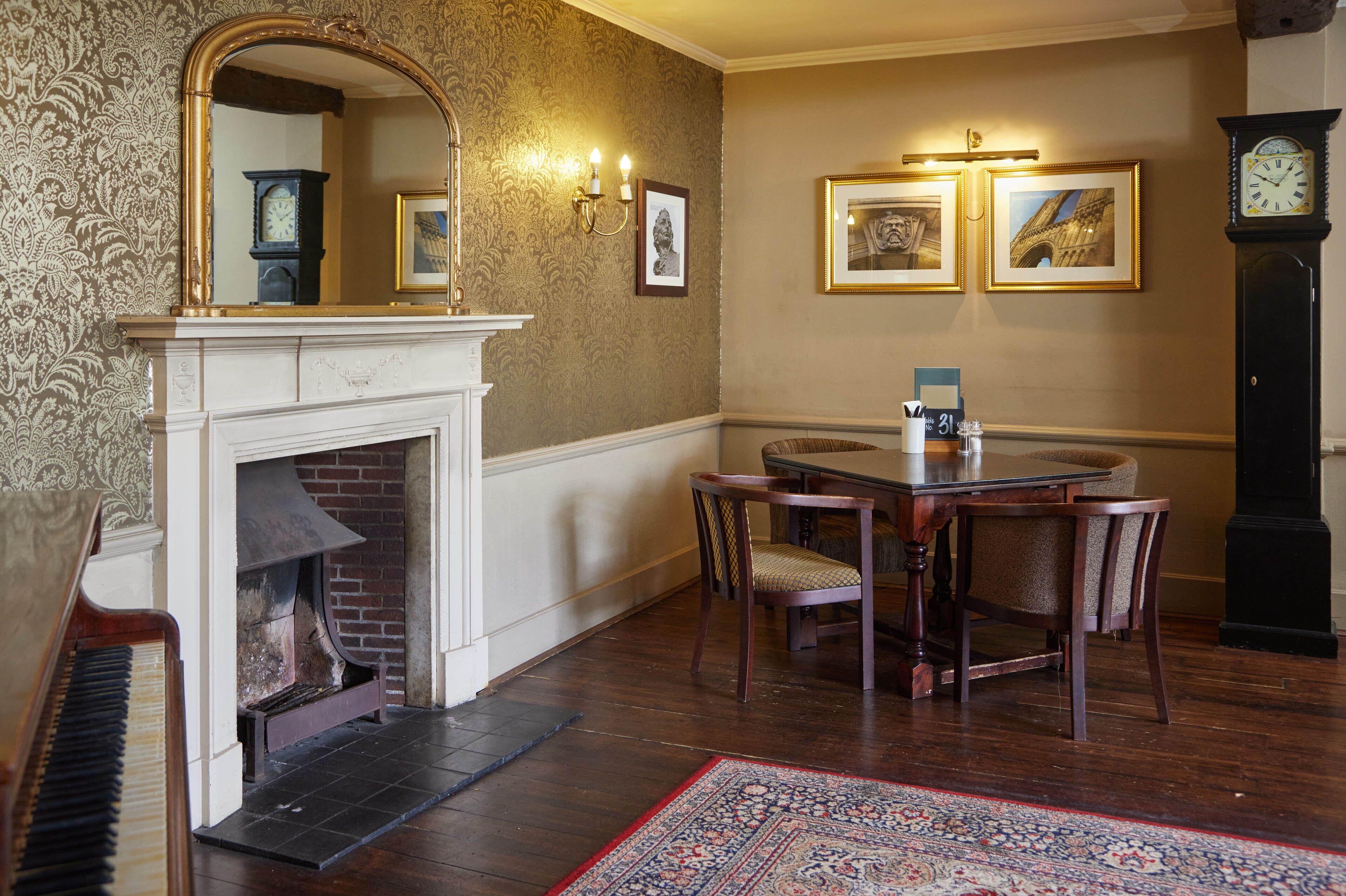A room with a fireplace, a dining table with chairs, a clock, and framed pictures on the wall.