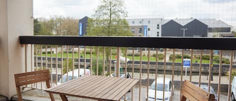 Un balcón con una mesa de madera y dos sillas, con vista a una calle con coches estacionados y edificios al fondo.