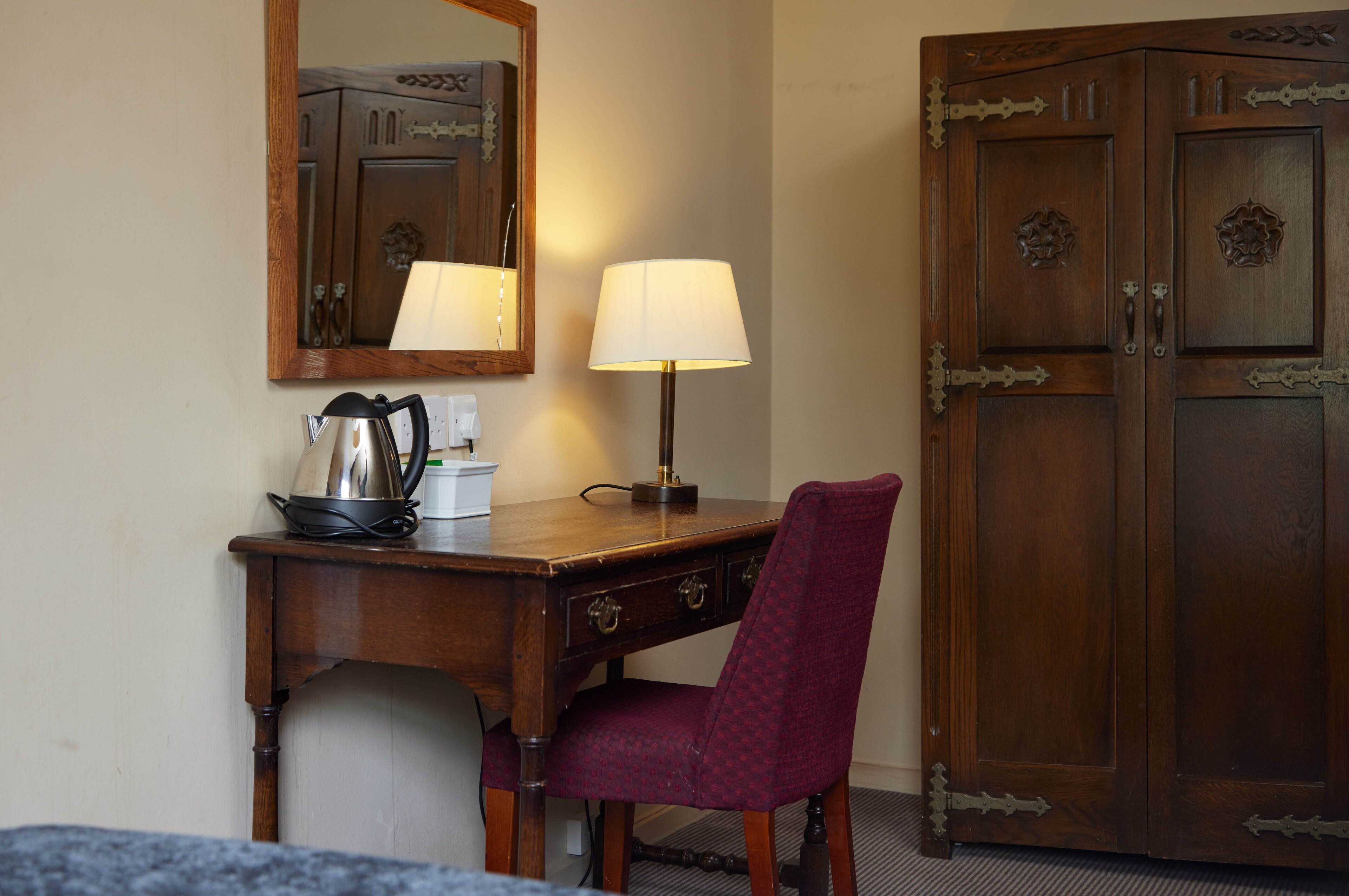 A wooden desk with a lamp, a chair, and a mirror reflecting a cabinet.