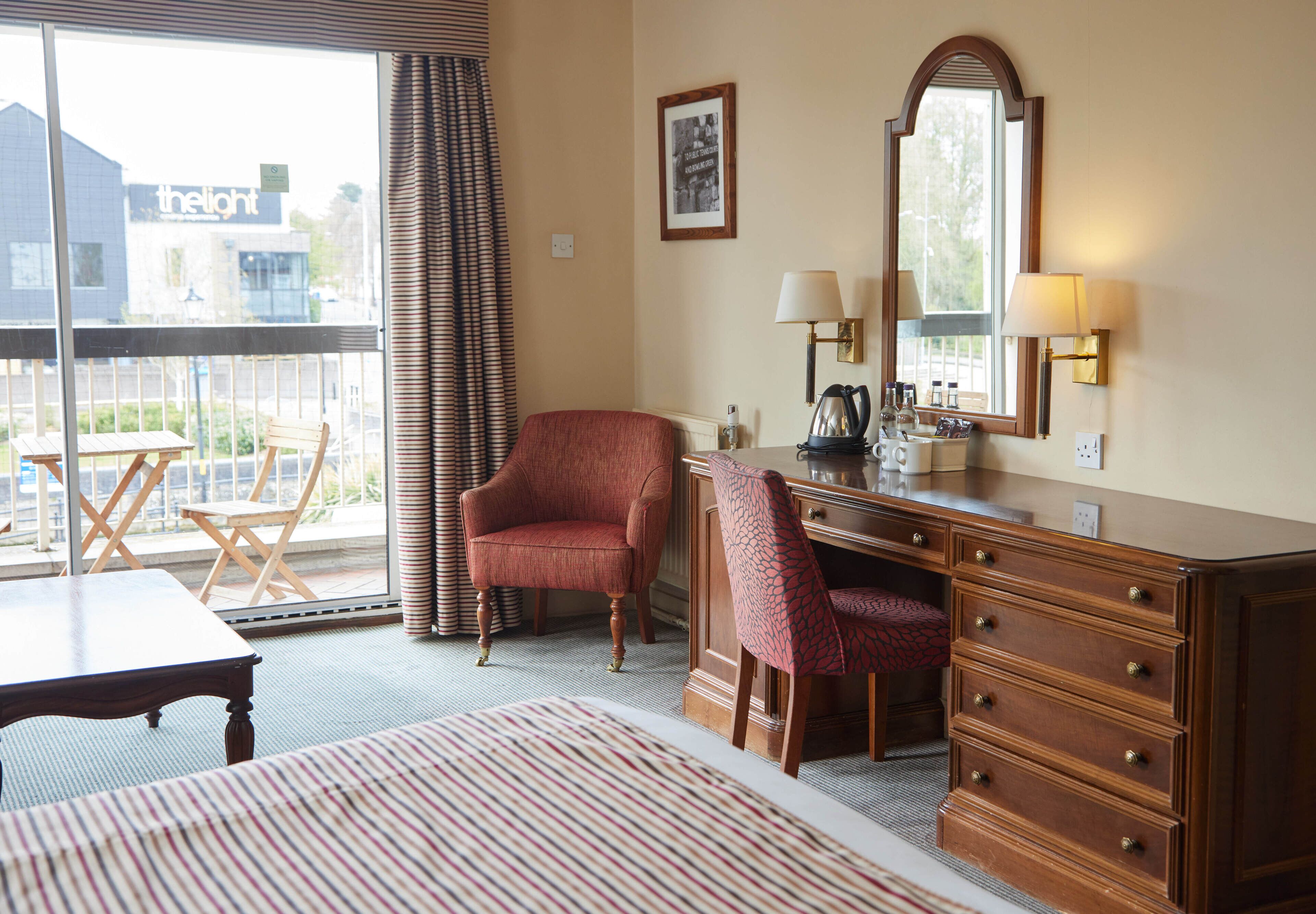 A hotel room with a bed, a desk, a chair, a dresser, a window with curtains, and a balcony with a table and chairs.