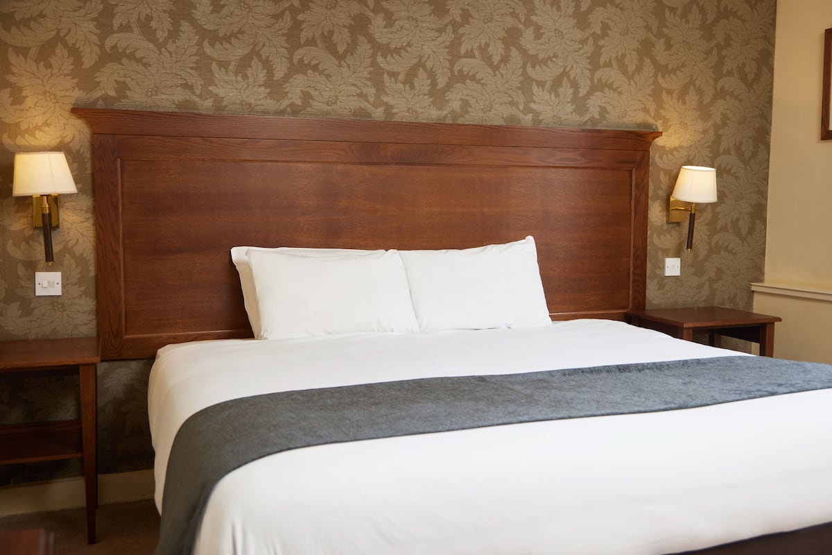 Una cama bien hecha con sábanas blancas y un plaid gris, flanqueada por dos mesitas de noche con lámparas, en una habitación con papel tapiz estampado y cabecera de madera.