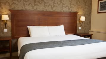 Una cama bien hecha con sábanas blancas y un plaid gris, flanqueada por dos mesitas de noche con lámparas, en una habitación con papel tapiz estampado y cabecera de madera.