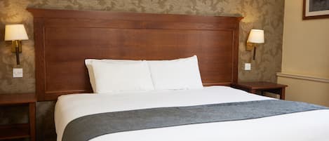 A neatly made bed with white linens and a gray throw, flanked by two nightstands with lamps, in a room with patterned wallpaper and a wooden headboard.