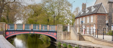 Un puente histórico con barandillas ornamentales cruza un río tranquilo, junto a un edificio tradicional de ladrillo con un techo a dos aguas.