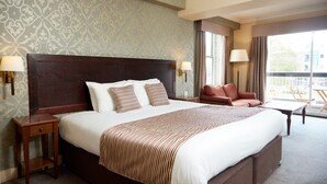 A hotel room with a large bed, two bedside lamps, a red armchair, and a view of the outdoors through a window.