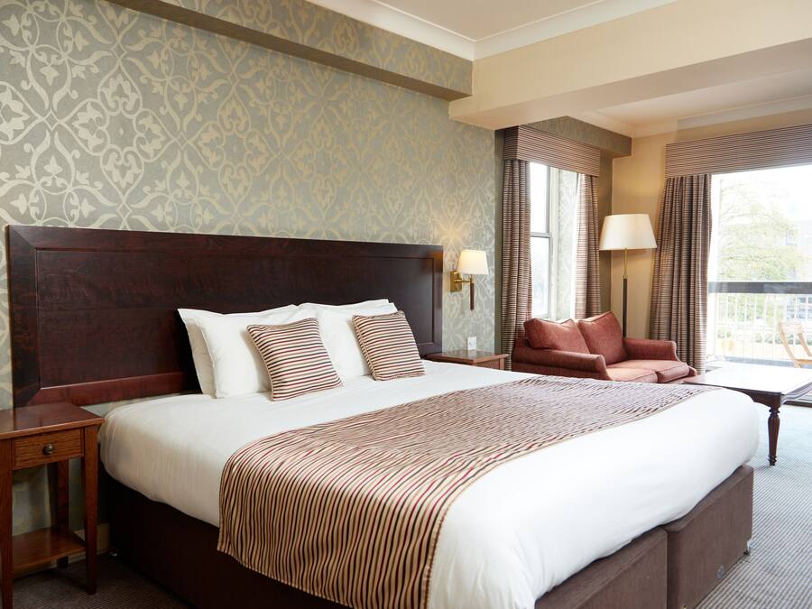 A hotel room with a large bed, two bedside lamps, a red armchair, and a view of the outdoors through a window.