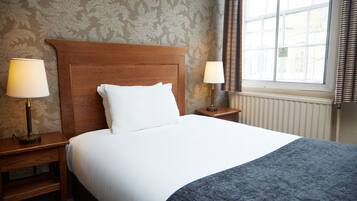 A neatly made bed with white linens and a blue throw blanket, flanked by two bedside tables with lamps, in a room with floral wallpaper and a window with curtains.