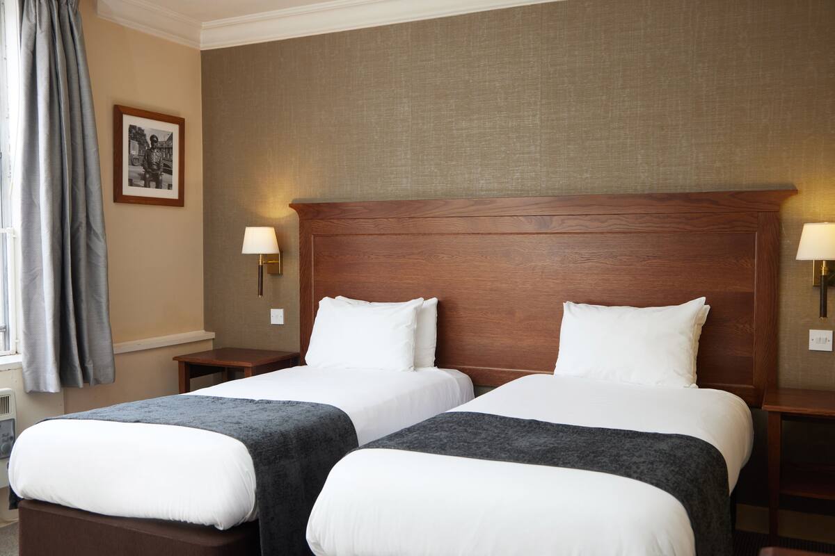 Habitación de hotel con dos camas, cabecera de madera, lámparas de noche, un cuadro en la pared y una ventana con cortinas.