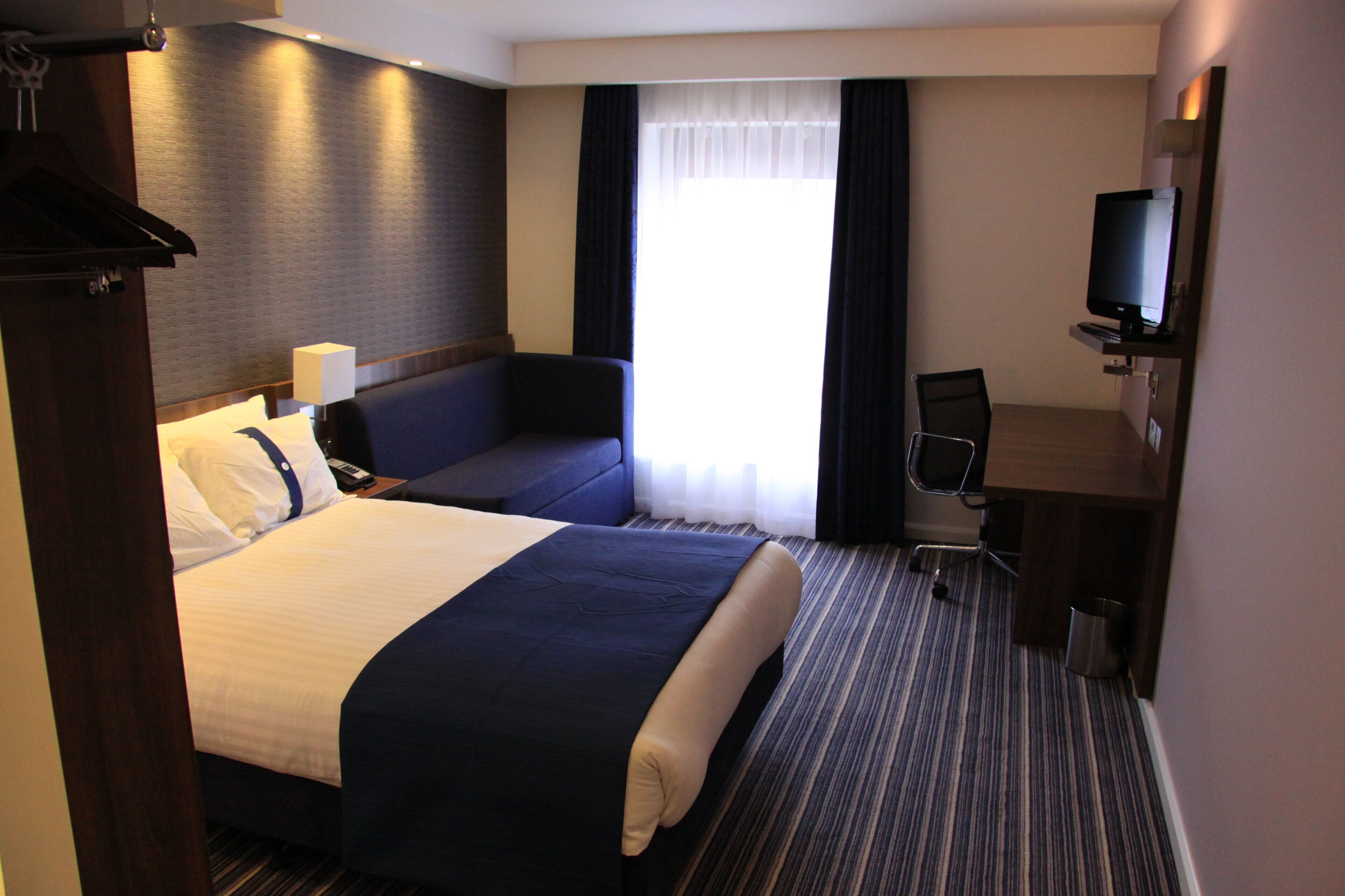 Una habitación de hotel con una cama grande, zona de estar, escritorio, televisor y ventana con cortinas.
