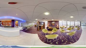 A modern hotel lobby with a reception desk, seating area, and a view of the city through large windows.