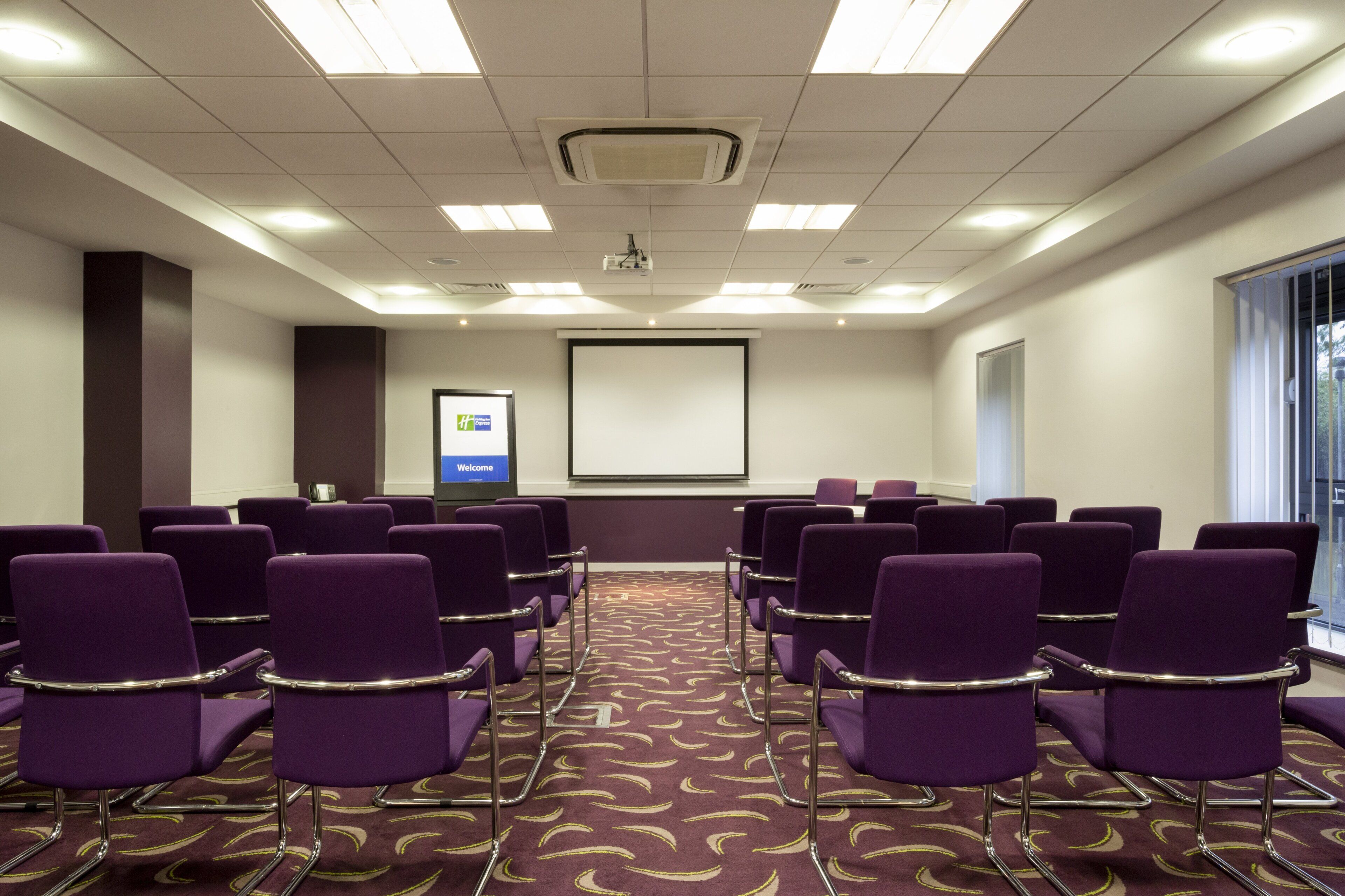A conference room with purple chairs arranged in rows, a projector screen, and a 'Welcome' sign.