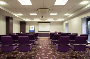 Un salón de conferencias con sillas moradas dispuestas en filas, una pantalla de proyector y un letrero que dice 'Welcome'.