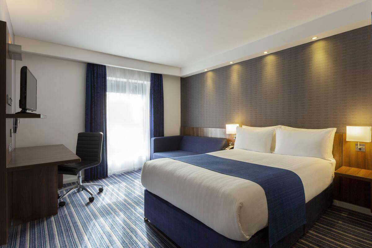 A hotel room with a large bed, a desk, a chair, and a nightstand with a lamp.