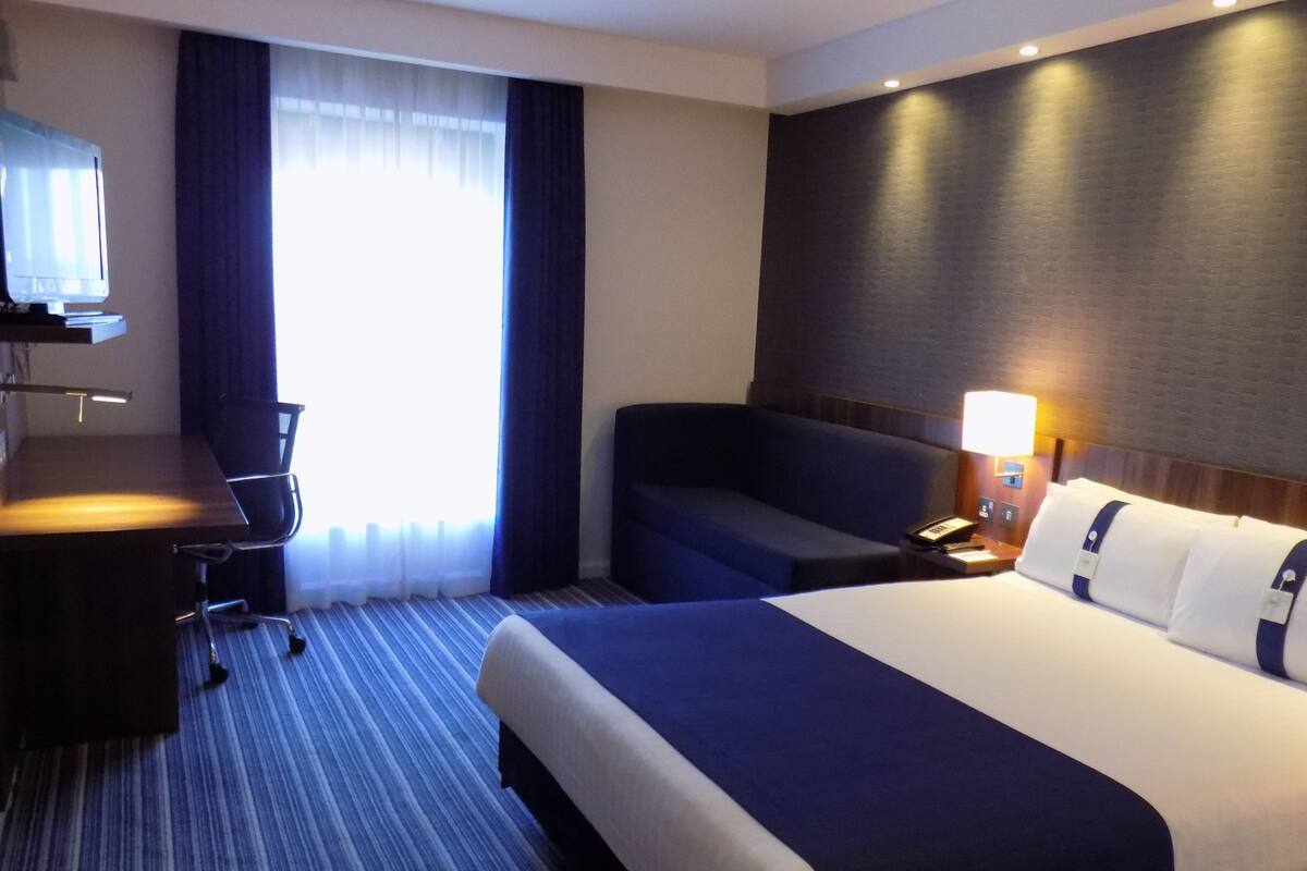 A hotel room with a bed, a desk, a chair, a TV, and a window with curtains.