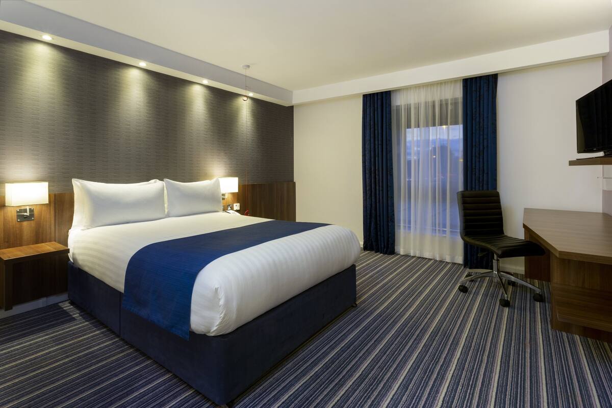 A hotel room with a large bed, a desk, a chair, a TV, and a window with curtains.