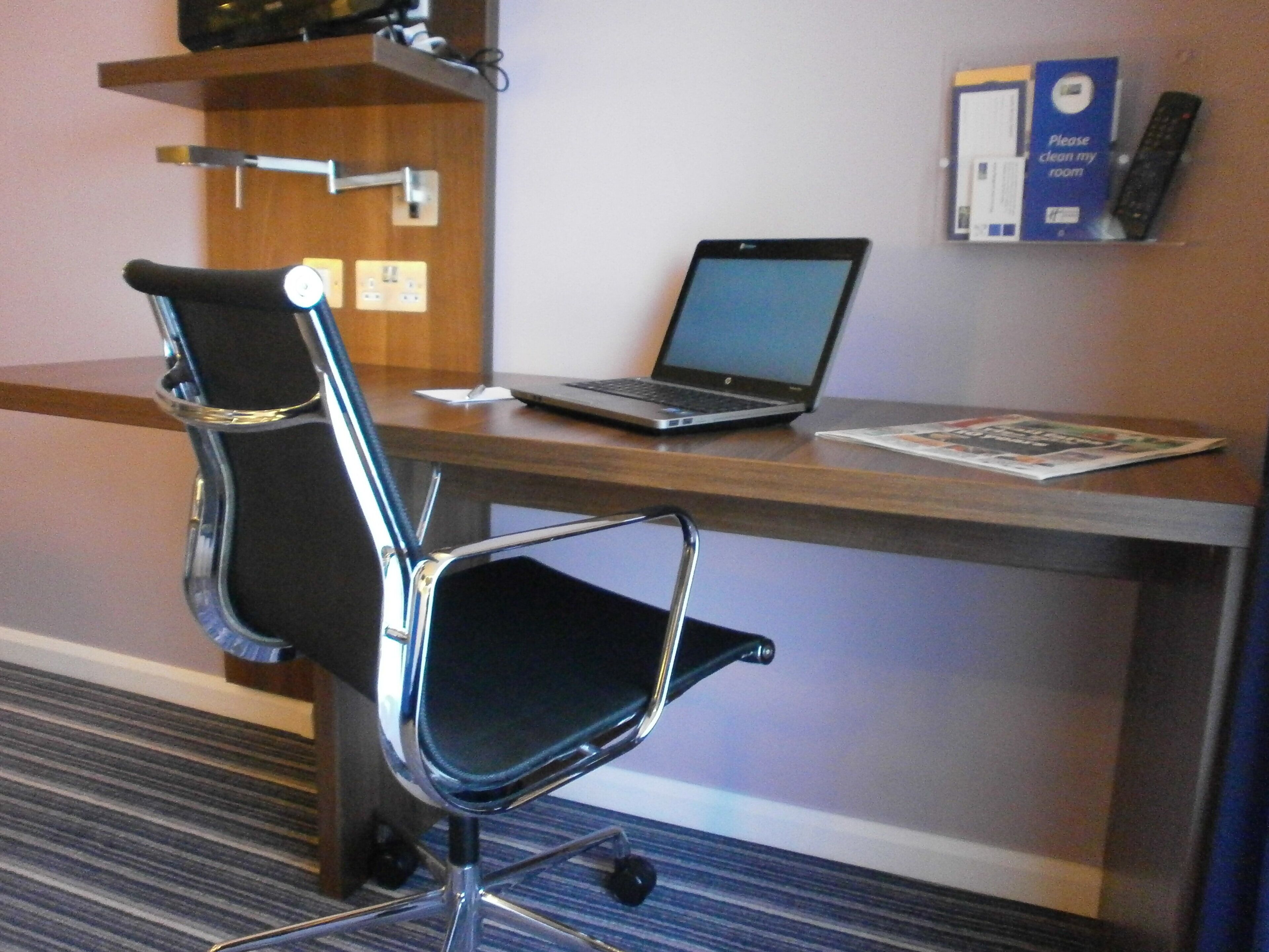 A hotel room with a desk, a laptop, a chair, and a wall-mounted shelf with a remote control and a 'Please clean my room' sign.