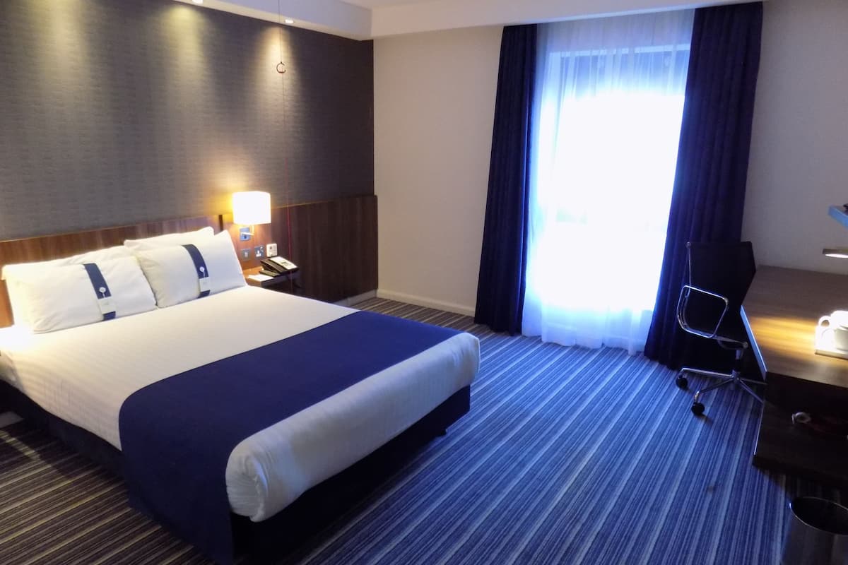 A hotel room with a large bed, a desk, a chair, a lamp, and a window with curtains.