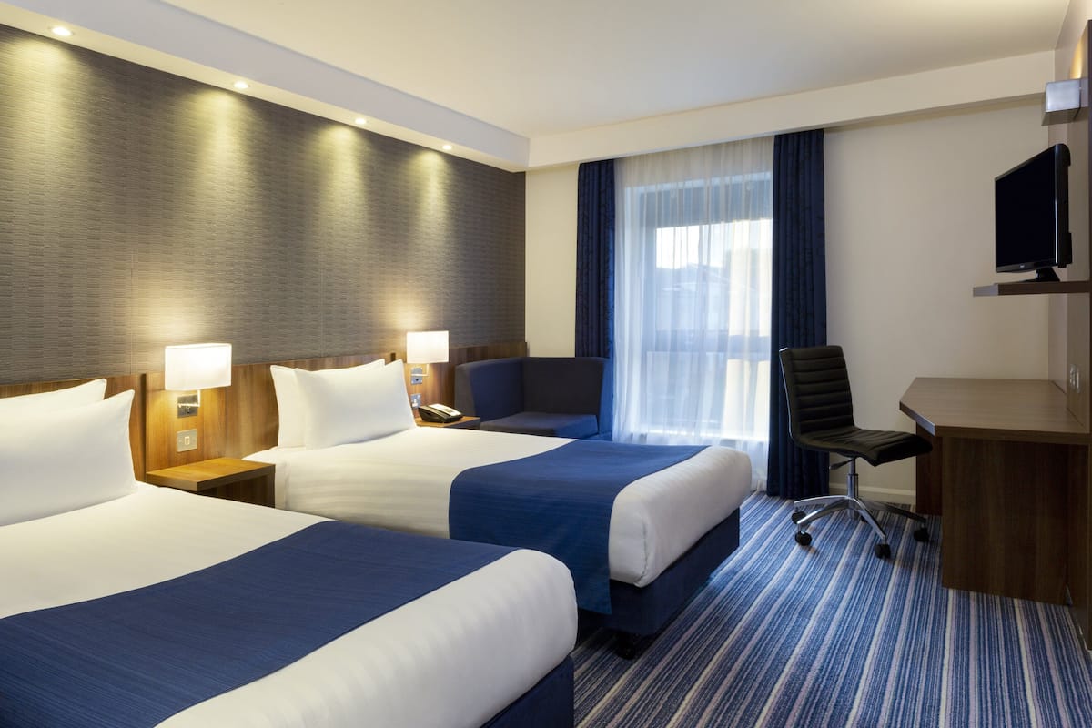 A hotel room with two beds, a desk, a chair, a television, and a large window with curtains.