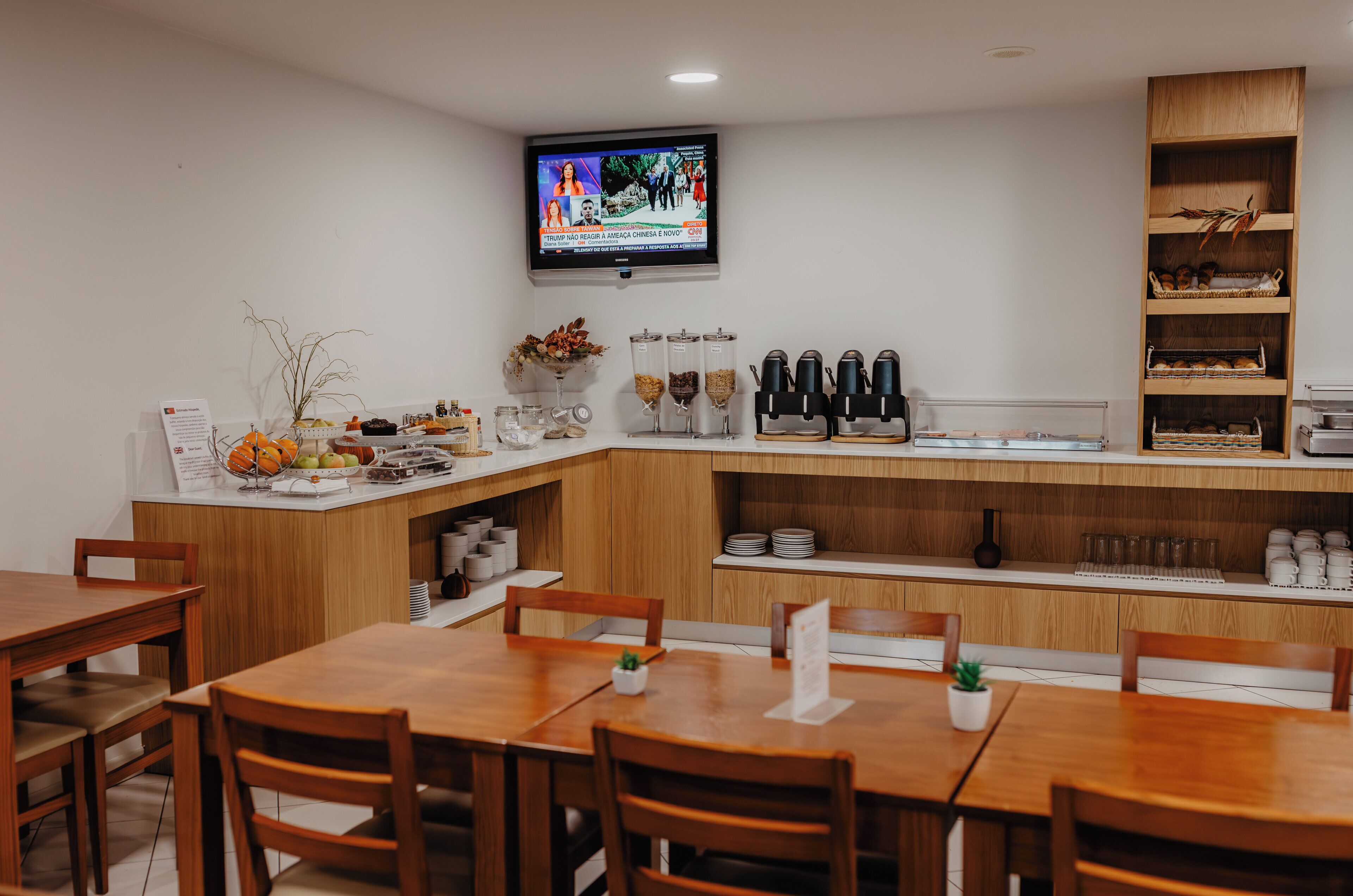 A modern dining area with wooden tables and chairs, a coffee station, and a television mounted on the wall.