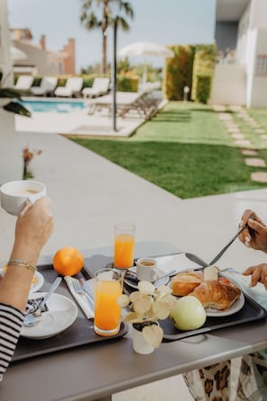 A breakfast tray with orange juice, a croissant, and an apple on a table with a pool view. - Hotel Sao Sebastiao de Boliqueime (Loulé)
