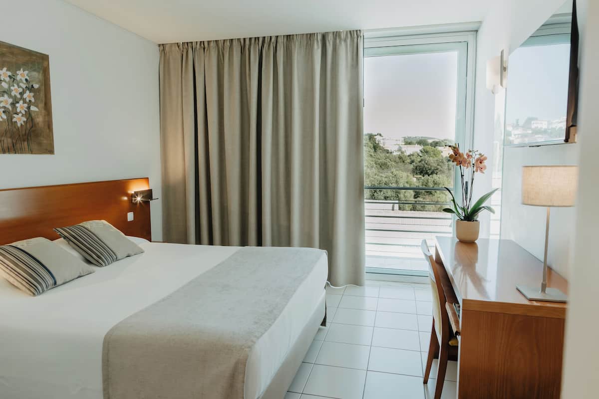 A hotel room with a bed, a desk, a chair, a window with curtains, and a potted plant.