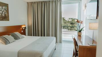 Habitación de hotel con cama, escritorio, silla, ventana con cortinas y una planta en maceta.