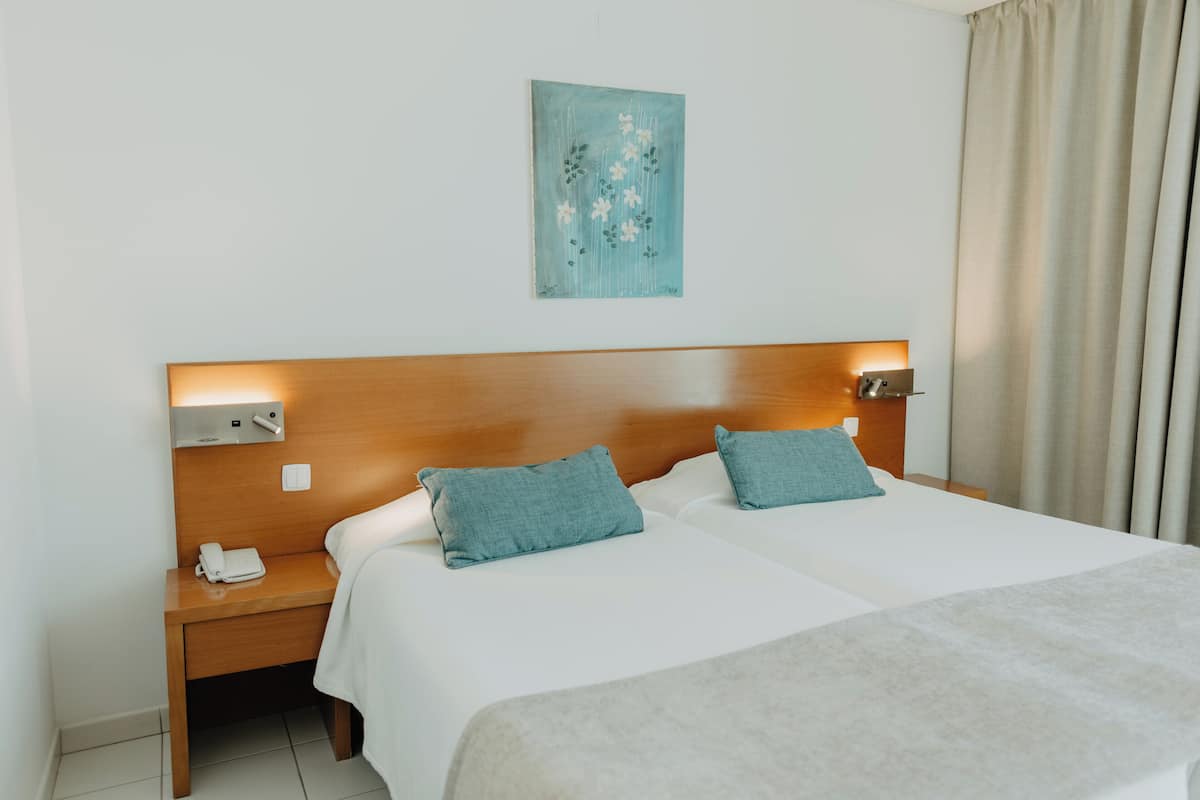A hotel room with a wooden headboard, two beds with teal pillows, a bedside table with a phone, and a painting on the wall.