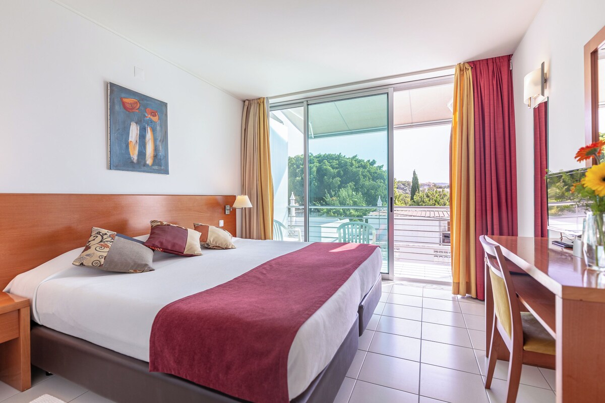 A hotel room with a large bed, a desk, and a balcony with a view of greenery.