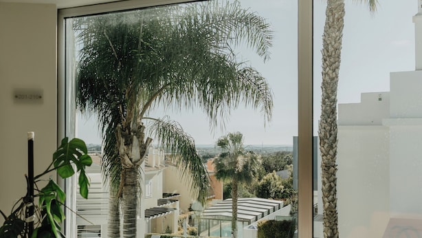A modern living room with a glass coffee table, a red sofa, and a view of a pool and palm trees. - Hotel Sao Sebastiao de Boliqueime (Loulé)